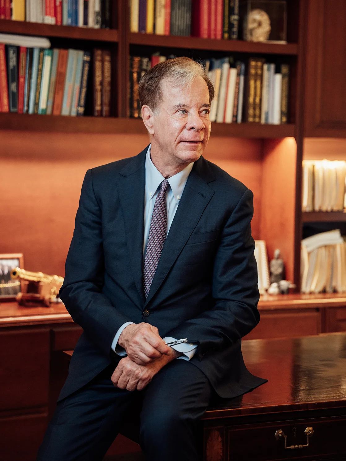 Dr Antell in navy suit sitting on desk in library office with bookshelf background - in New York, NY