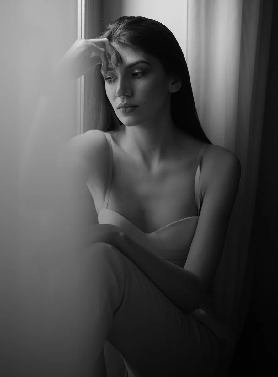 Woman in contemplative pose by window in black and white portrait with natural lighting and artistic shadows - breast in New York, NY