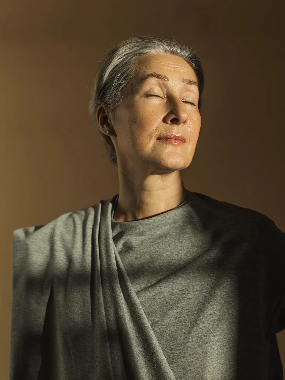 Serene woman with short gray hair in sunlight wearing gray t-shirt with eyes closed in peaceful contemplation - in New York, NY