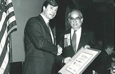 Two men in suits shaking hands during formal ceremony with American flag and framed certificate visible