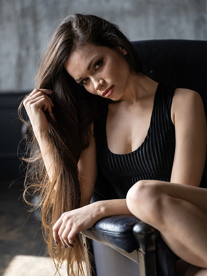 Woman in black ribbed dress posing with long brown hair against grey textured background - breast lift in New York, NY