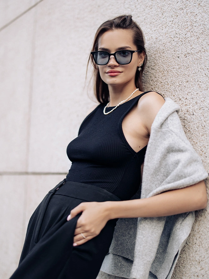 Pregnant woman in black dress and sunglasses leaning against white wall wearing grey cardigan - breast reduction in New York, NY