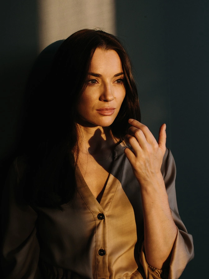 Woman with long dark hair in beige cardigan gesturing with hands in natural sunlight - facial implants in New York, NY