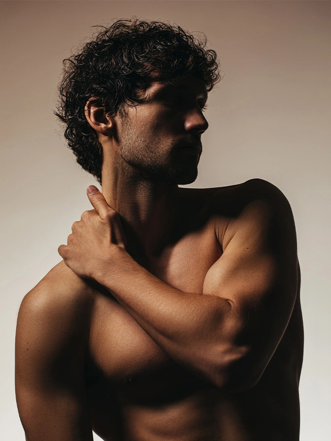 Shirtless man with curly dark hair touching shoulder in profile against neutral background - gynecomastia in New York, NY