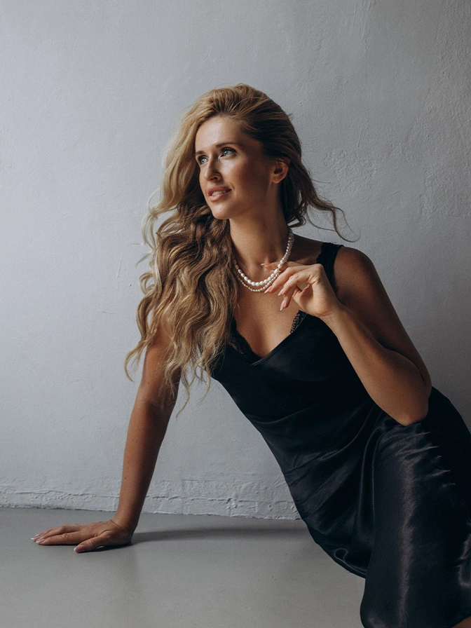 Elegant woman in black evening dress with long blonde hair posing against minimalist gray wall - lipoma removal in New York, NY