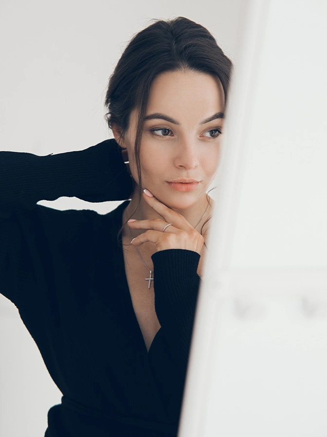 Woman in black turtleneck with dark hair pulled back, hand under chin in thoughtful pose against white background - mole removal in New York, NY
