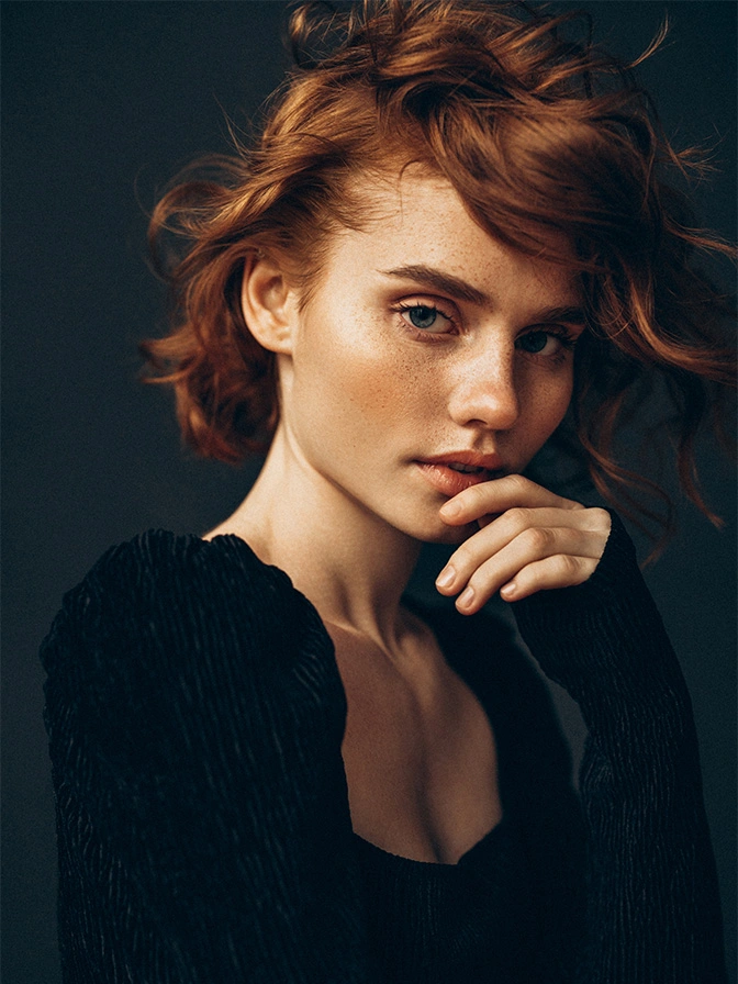Woman with short wavy auburn hair in black top against dark background, hand near face in thoughtful pose - non surgical rhinoplasty in New York, NY