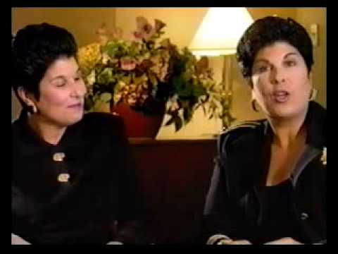 Two women in black attire seated during a press interview with flowers and lamp in background