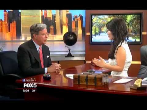 Two news anchors in professional attire conducting interview at Fox 5 studio desk with colorful backdrop and globe